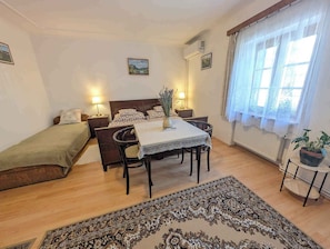 A bedroom with a bed, a small table with chairs, a bedside table with lamps, a window with curtains, and a potted plant.