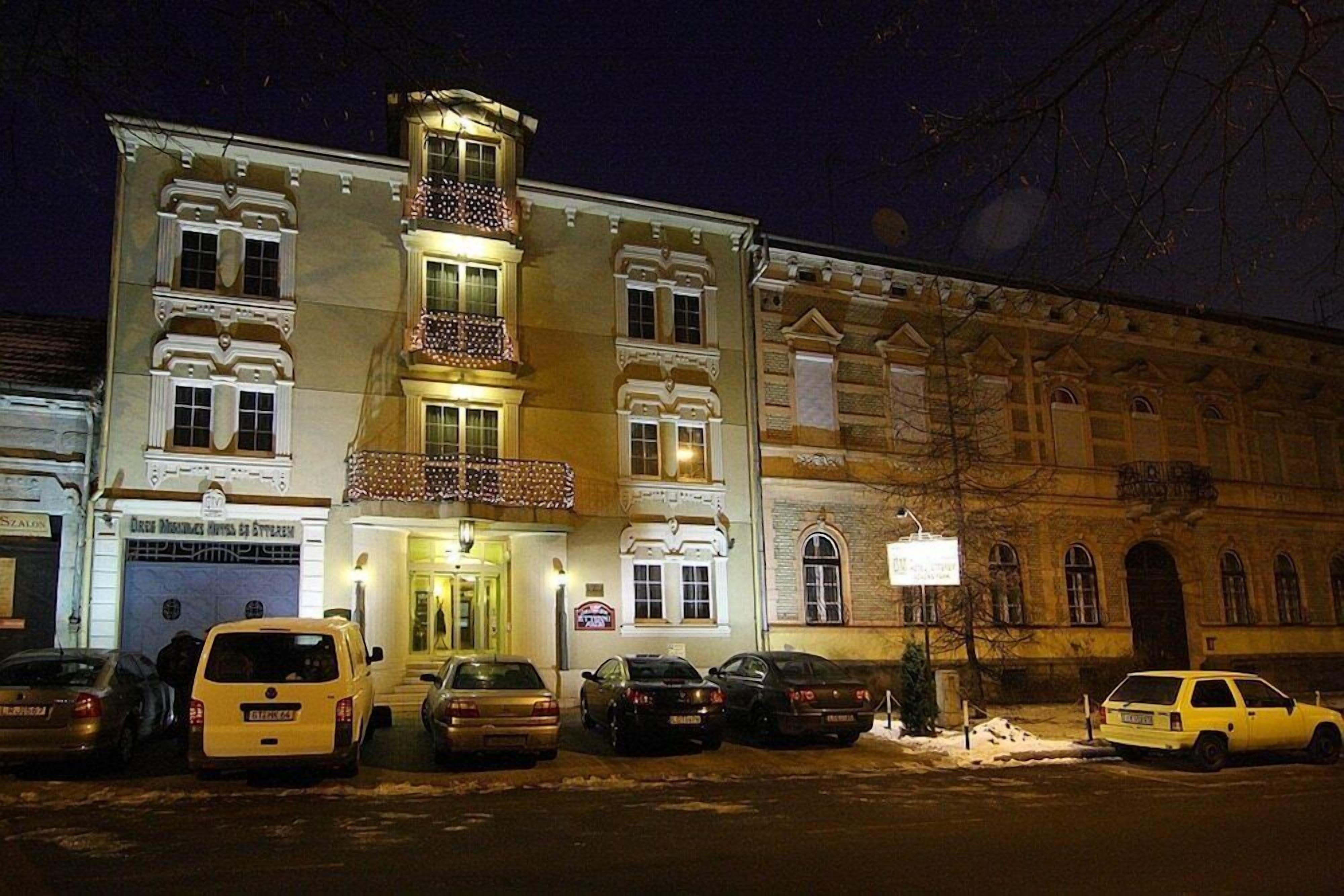 A multi-story building with a central balcony, a ground entrance, and several parked cars.