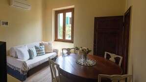 Un comedor con una mesa redonda de madera, sillas blancas, un jarrón con flores, un ventanal con vistas y una puerta de madera.