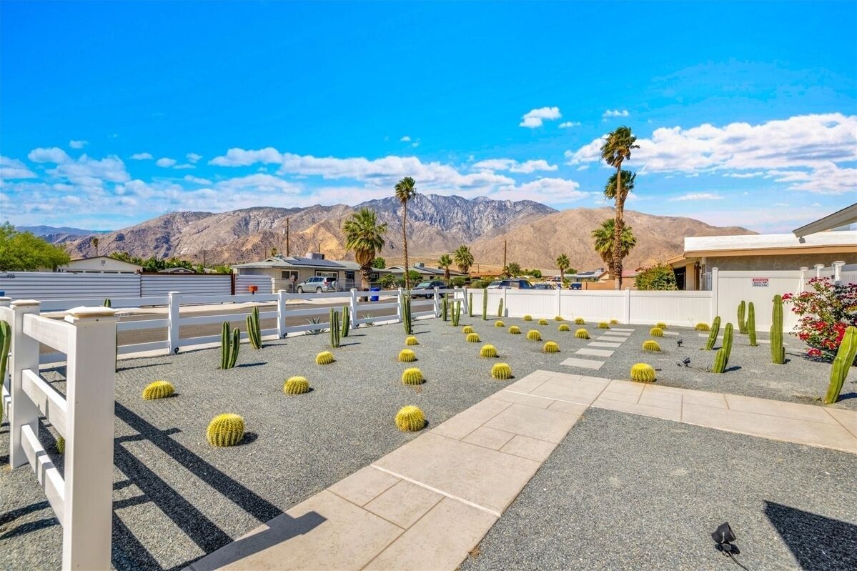 A parking area with cacti and palm trees, a white fence, and mountains in the background.
