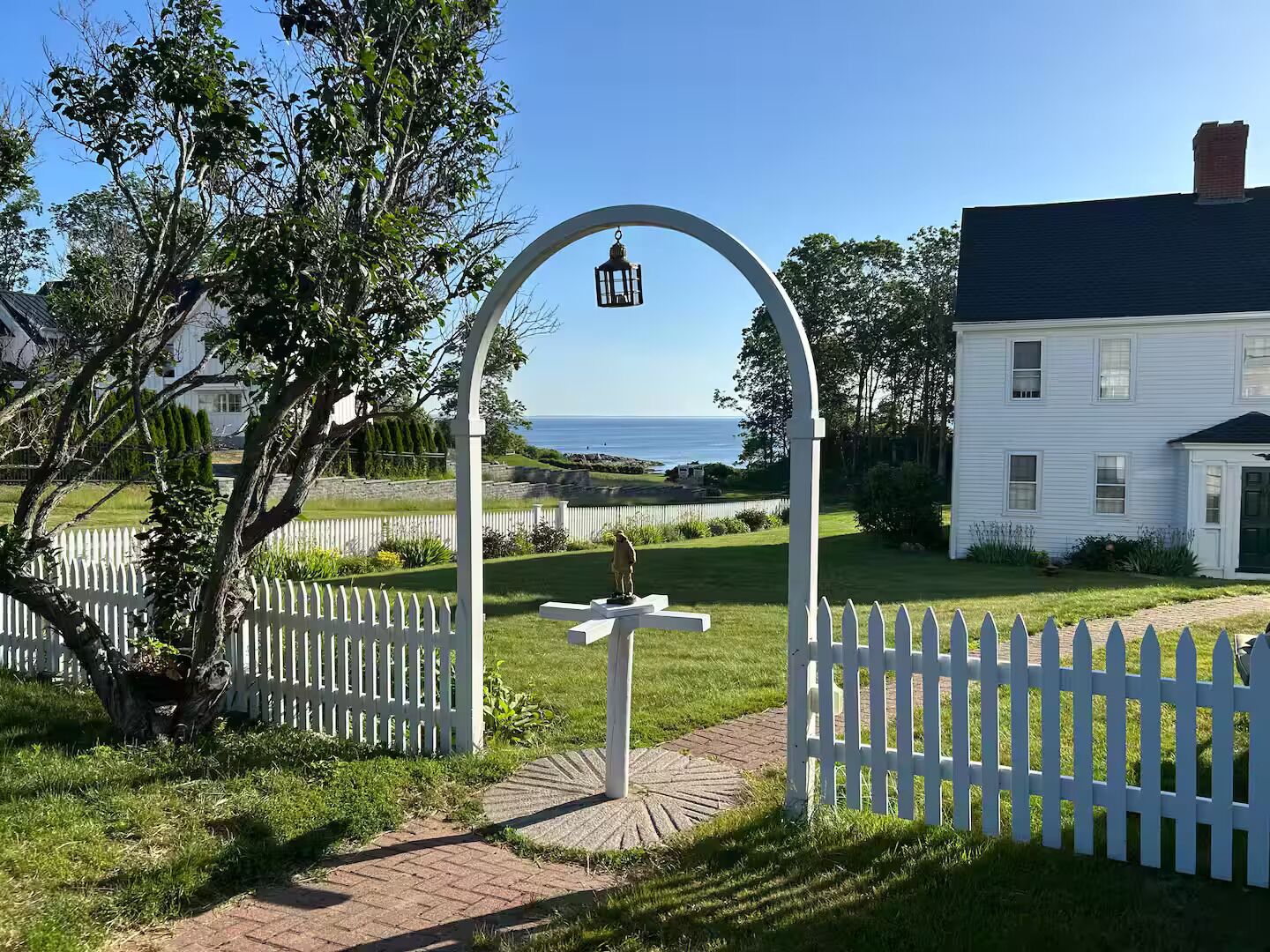 A white house with a black roof, a white picket fence, and a view of the ocean.