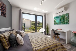 A modern bedroom with a large bed, a desk, and a view of greenery outside.