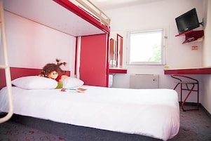 Una cama individual con colchón blanco, cabecera roja, mesita de noche pequeña, televisor montado en la pared y ventana con luz natural.