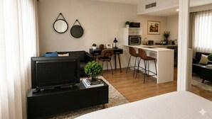 Ein modernes Wohnzimmer mit Fernseher, einem kleinen Couchtisch mit Büchern und einer Pflanze, einem Essbereich mit Stühlen, einer Küche mit Mikrowelle und Kühlschrank sowie einem großen Fenster mit Vorhängen.