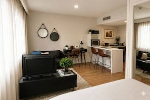 Ein modernes Wohnzimmer mit Fernseher, einem kleinen Couchtisch mit Büchern und einer Pflanze, einem Essbereich mit Stühlen, einer Küche mit Mikrowelle und Kühlschrank sowie einem großen Fenster mit Vorhängen.
