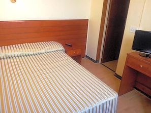 Habitación de hotel con una cama, cabecero de madera, televisor sobre un soporte de madera y una puerta que da a otra habitación.