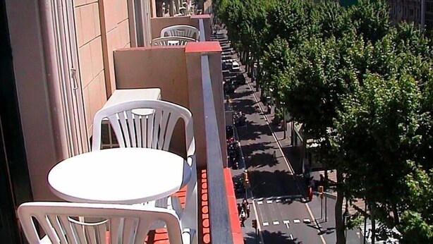 A balcony with white plastic chairs and a table, overlooking a street lined with trees and buildings. - Toledano (Barcelona)