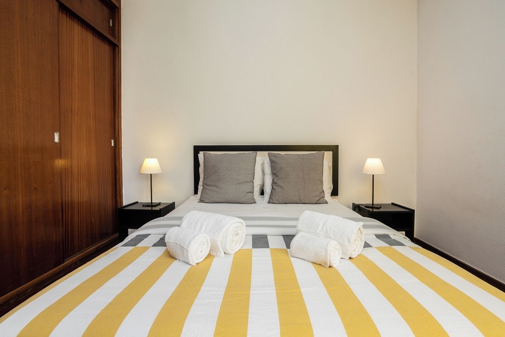 Ein Hotelzimmer mit gestreifter Bettdecke, zwei Kissen, zwei Nachttischen mit Lampen und einem Holzschrank.