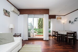 Ein Zimmer mit einem weißen Sofa, einem Esstisch mit Stühlen, Holzboden, Deckenventilator und Blick auf eine grüne Außenanlage.