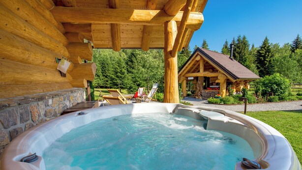 Un jacuzzi bajo una pérgola de madera con un muro de piedra, rodeado por un jardín y una cabaña.