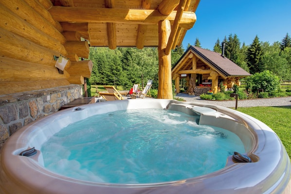 Un bain à remous sous une pergola en bois, adossé à un mur de pierre, entouré d’un jardin et d’un chalet.