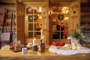 Une pièce chaleureuse aux murs en bois, une table décorée de bougies, un sapin de Noël et une vue sur un intérieur éclairé d’une lumière douce et accueillante.