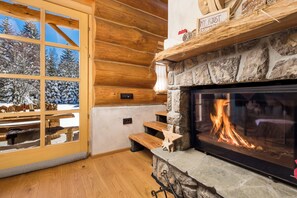 A log cabin interior with a stone fireplace, wooden benches, and a view of snow-covered trees outside.
