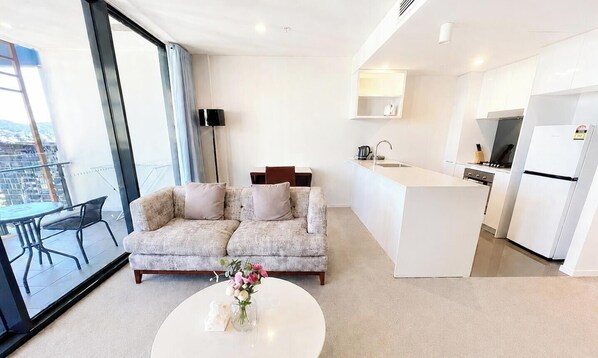 A modern living room with a sofa, a coffee table with flowers, a dining table, and a kitchen area with white cabinets and appliances. - Comfortable 2BR w/ Pool, Gym and BBQ | Free Parking (South Brisbane)