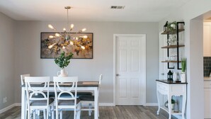 Un coin repas avec une table et des chaises blanches, un lustre moderne et un tableau abstrait accroché au mur.