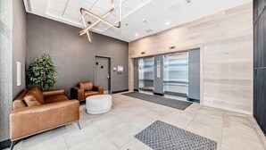 Um lobby com sofá de couro marrom, um banquinho branco, luminária de teto geométrica e dois elevadores.