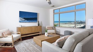 Ein modernes Wohnzimmer mit großem Fenster, Deckenventilator, Holztisch und Blick auf den Strand.
