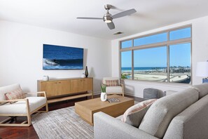 A modern living room with a large window, a ceiling fan, a wooden coffee table, and a view of the beach. - Ocean & Marina View Condo with Balcony, Pool, Hot Tub & Gym (Oceanside)