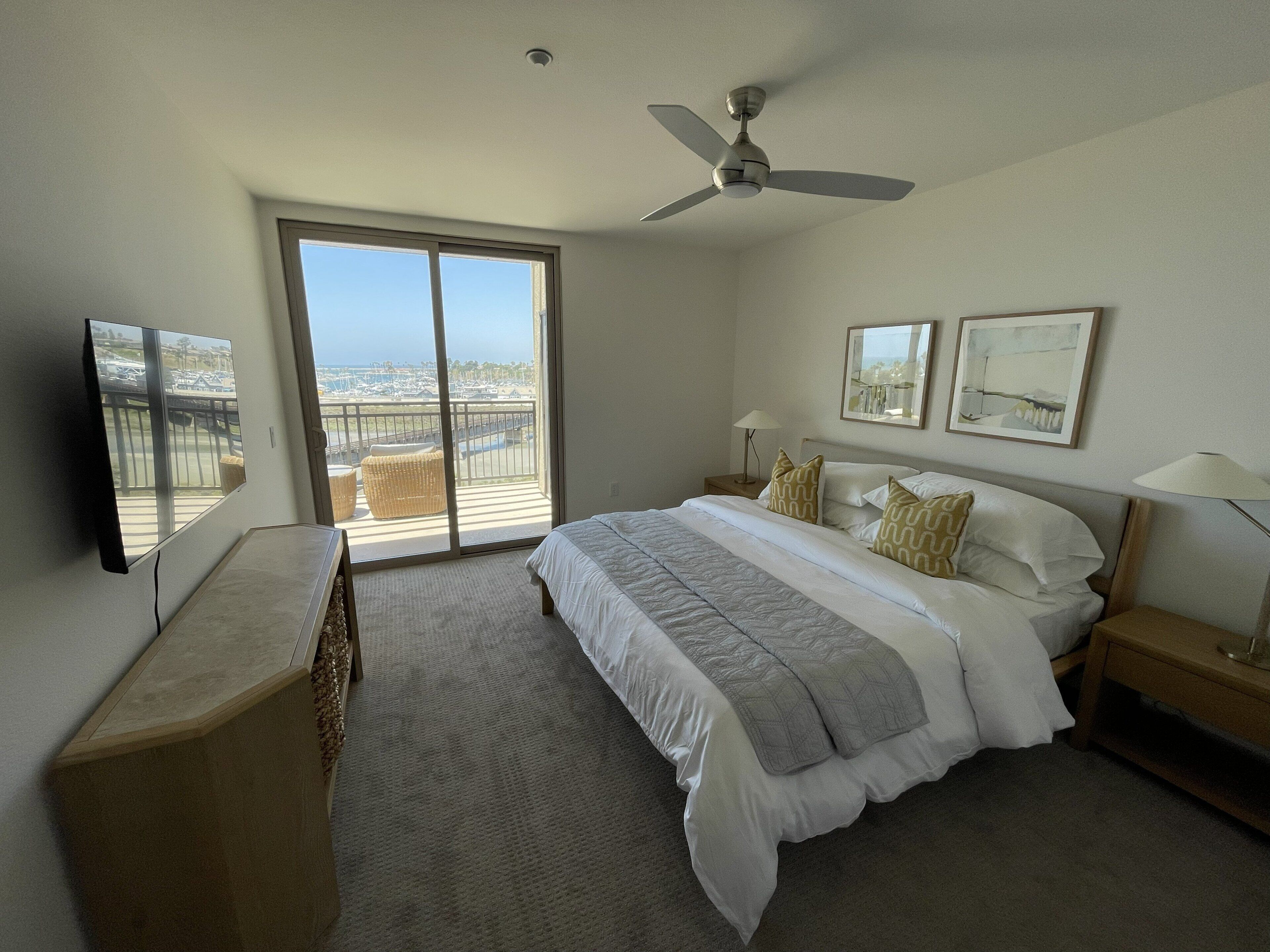 Habitación de hotel con cama, ventilador de techo, televisión, balcón con vistas y obras de arte en las paredes.