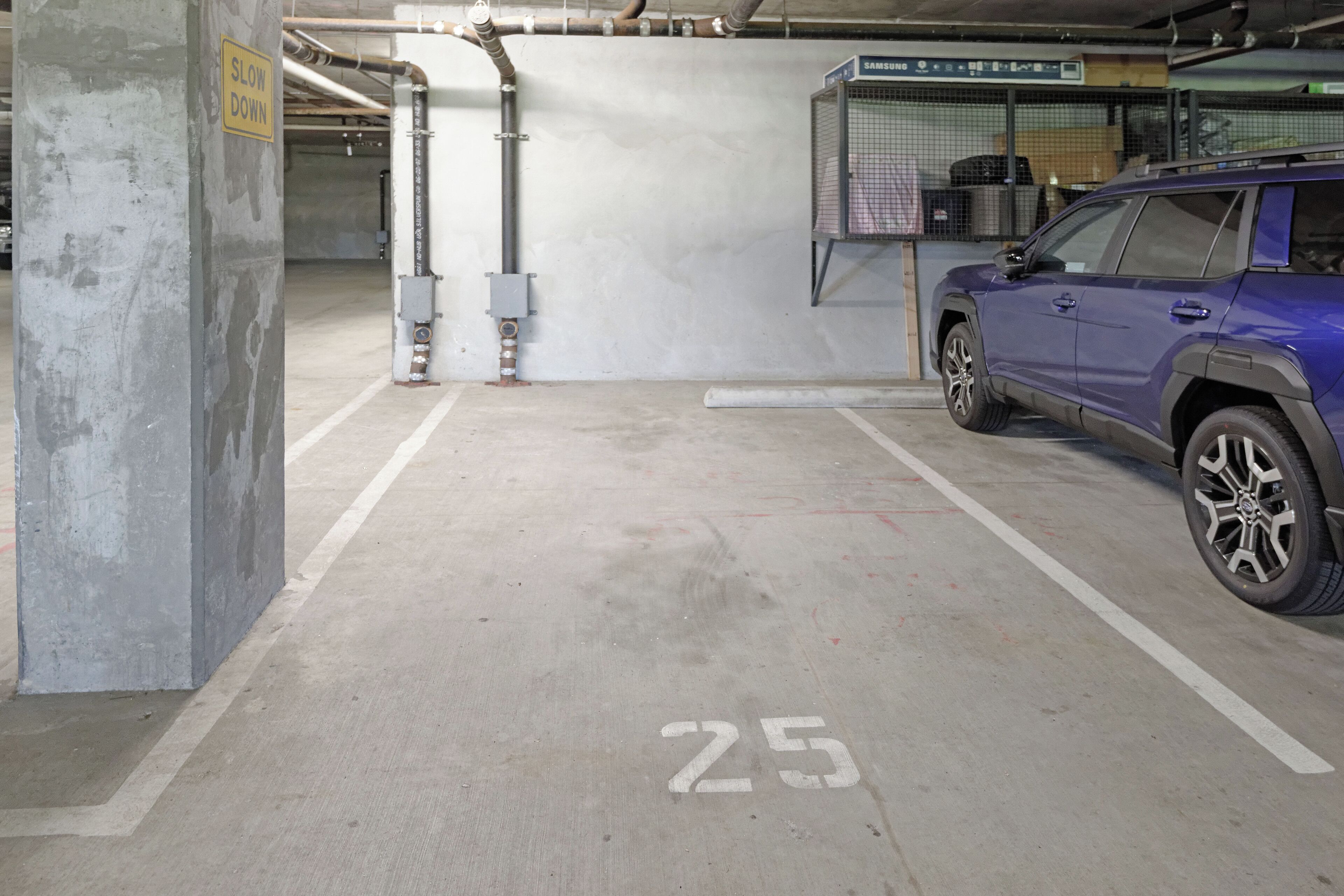 Un parking avec un panneau indiquant « RALENTISSEZ », un véhicule stationné sur la place numéro 25, et une pièce technique contenant un chauffe-eau.