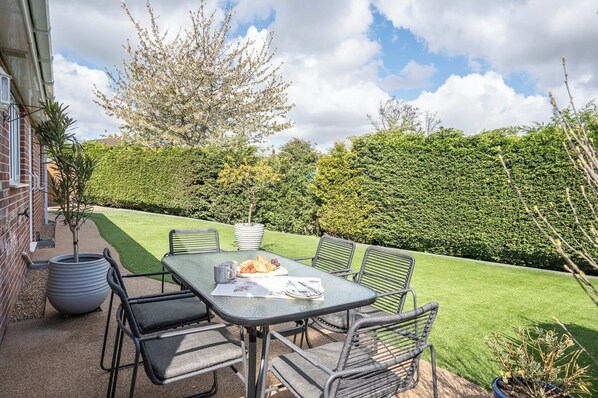 Eine Terrass mit Tisch, Stühlen, einer Kübelpflanze und einer Hecke im Hintergrund.