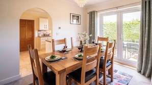 Un coin repas avec une table en bois, des chaises, un lustre et une vue sur un jardin, accessible par une porte-fenêtre coulissante.