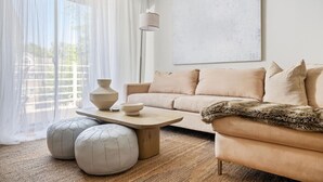 A living room with a sofa, ottomans, a coffee table, a lamp, and a large window with sheer curtains. - Light Filled 2BR w/ Balcony & 2 Free Parking (Arlington)