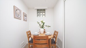 Uma sala de jantar com uma mesa redonda, quatro cadeiras de madeira, pratos brancos, um vaso com flores e duas obras de arte emolduradas na parede.