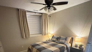 A bedroom with a bed, a ceiling fan, a window with curtains, a nightstand with a lamp, and a small plant.