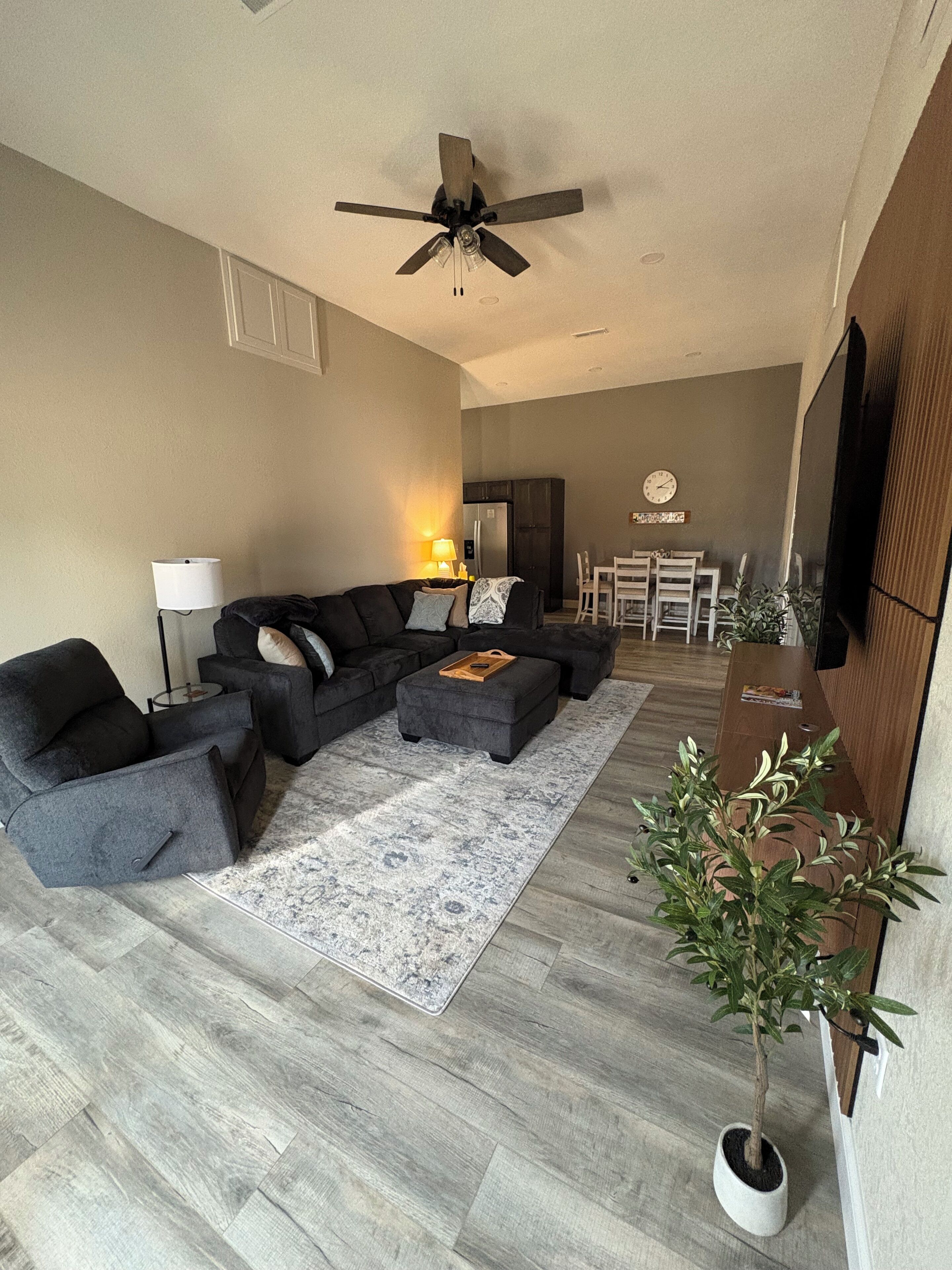 A modern living room with a sectional sofa, a coffee table, a dining area with chairs, a TV, and a ceiling fan.