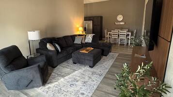 A modern living room with a sectional sofa, a coffee table, a dining area with chairs, a TV, and a ceiling fan.