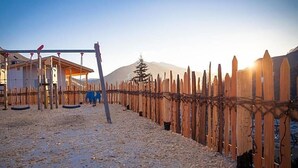 A playground with swings and a climbing structure, surrounded by a wooden fence and a mountain in the background. - Berghotel Schopfenhof (Dobbiaco)