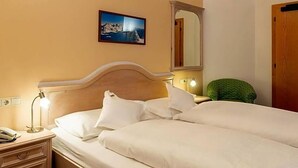 A hotel room with a bed, bedside lamp, a green chair, a mirror, and a painting on the wall. - Berghotel Schopfenhof (Dobbiaco)