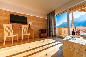 A wooden interior with a TV, two chairs, a table, a lamp, and a view of mountains. - Berghotel Schopfenhof (Dobbiaco)