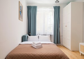 Ein ordentlich bezogenes Bett mit einer gesteppten Decke, ein Stapel gefalteter Handtücher und ein Fenster mit durchsichtigen Vorhängen.
