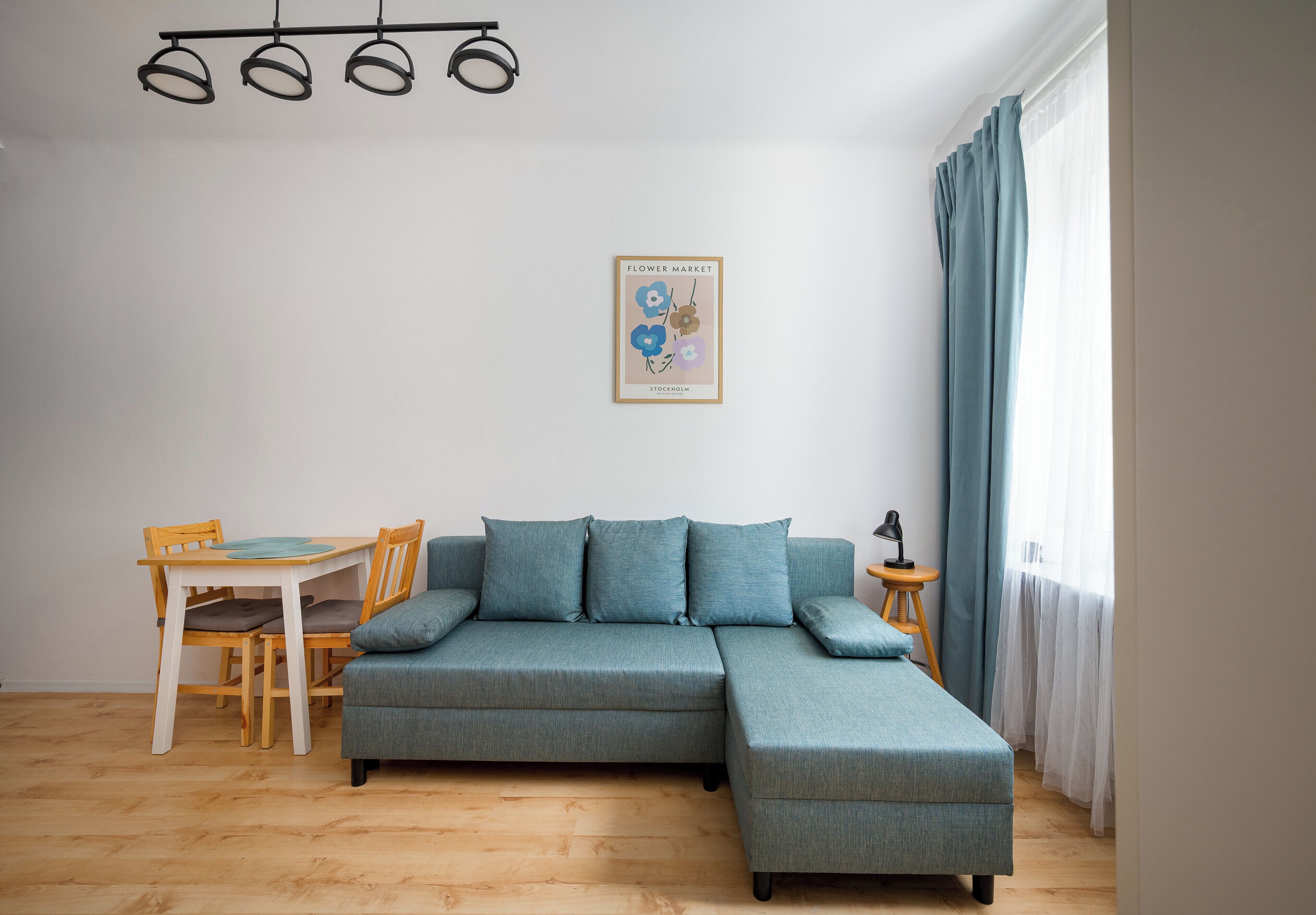 A living room with a sectional sofa, a dining table with chairs, a small side table, and a wall-mounted light fixture.