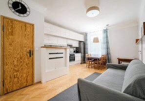 Eine moderne Wohnung mit Holztür, weißen Schränken, einer Küche mit Kühlschrank, einem Essbereich mit Tisch und Stühlen sowie einem grauen Sofa.