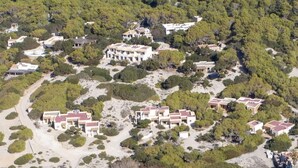 An aerial view of a coastal area with a cluster of buildings surrounded by dense greenery and a sandy beach. - Las Dunas Apt (Formentera)