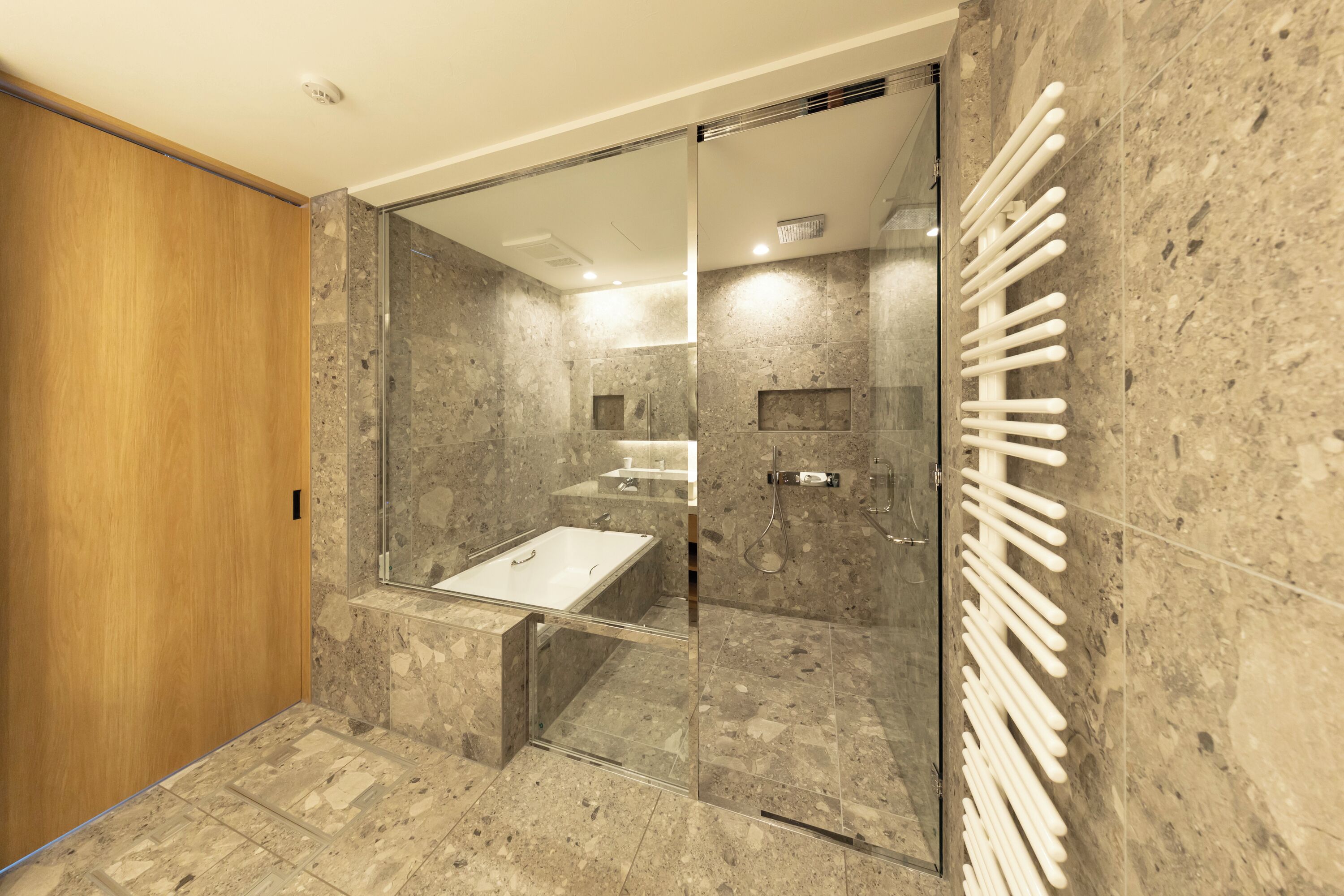 A modern bathroom with a glass shower enclosure, a wooden door, and a marble-tiled floor.