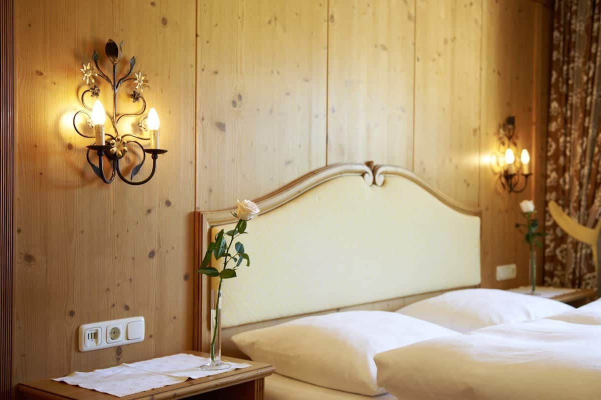 Ett hotellrum med en väggpanel av trä, en vit säng med ett böjt sänggavel, ett sängbord med en lampa och en vas med blommor samt ett eluttag.