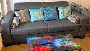 Un canapé gris avec trois coussins, une peinture abstraite aux couleurs vives accrochée au-dessus, et une petite table à la surface réfléchissante.
