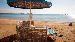 A beach umbrella, a wicker chair, and a striped lounge chair on a sandy beach with boats in the background. - Grand Resort (Hurghada)