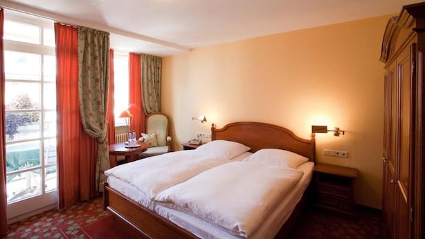 A hotel room with a double bed, a chair, a desk with a lamp, a nightstand, a wardrobe, and a window with curtains. - Karpfen (Eberbach)