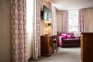A room with a wooden desk, a purple sofa, patterned curtains, and a TV mounted on the wall. - Karpfen (Eberbach)