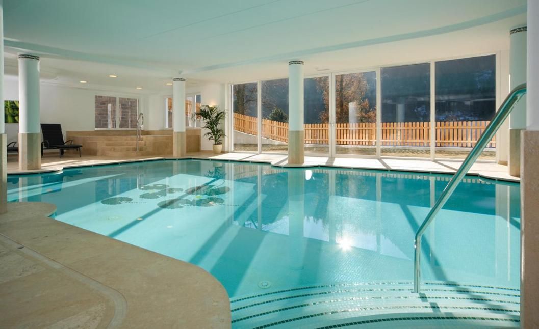 An indoor swimming pool with clear glass walls, a wooden deck outside, and a lounge chair inside.