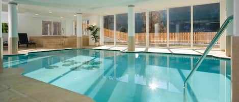 An indoor swimming pool with clear glass walls, a wooden deck outside, and a lounge chair inside.