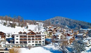 Uma aldeia montanhosa coberta de neve com edifícios estilo chalé, uma pista de esqui e um céu azul límpido.