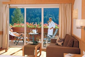 A room with a sofa, a table with a glass top, a chair, and a balcony with a view of a forest and flowers. - Grones (Ortisei)