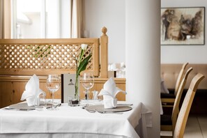 Mesa de jantar posta com toalha branca, copos de vinho, um vaso com flor e suporte para menu.