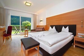 Habitació d'hotel amb un llit gran, un panell de capçal de fusta, una taula de nit, una cadira i vista a un paisatge verd a través de la finestra.